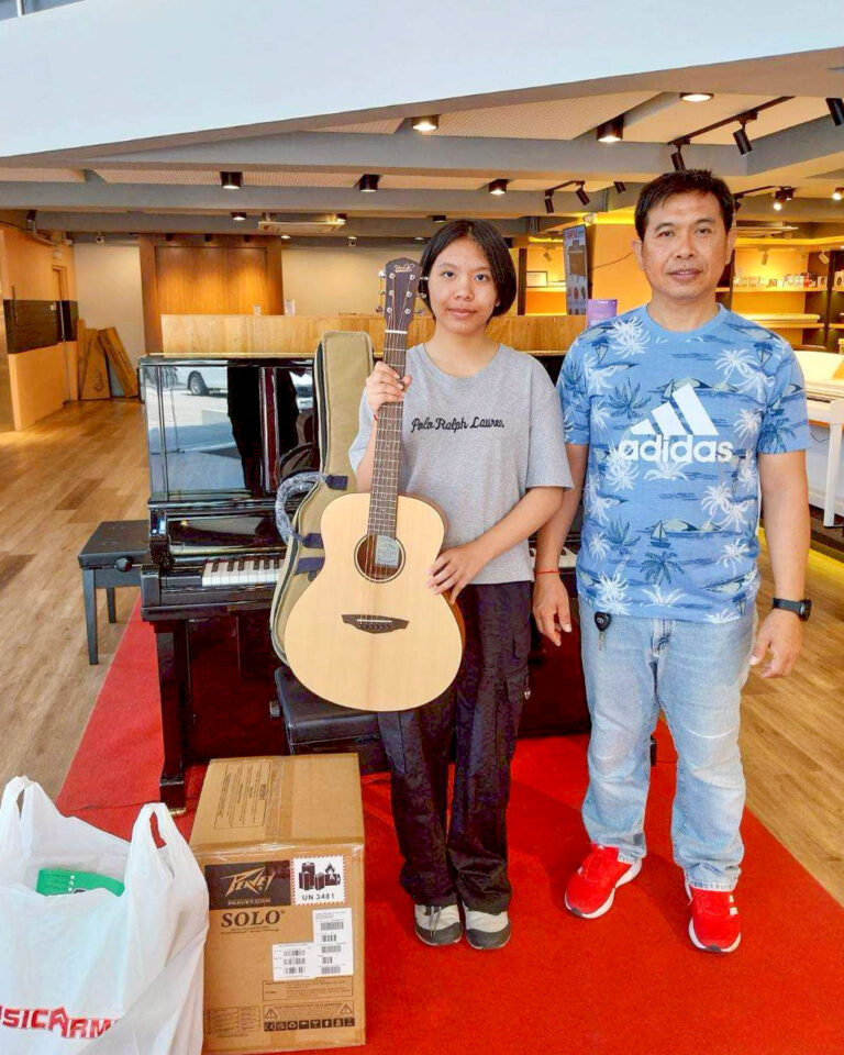 Young woman holds an acoustic guitar beside a man in a blue Adidas shirt inside a music store.