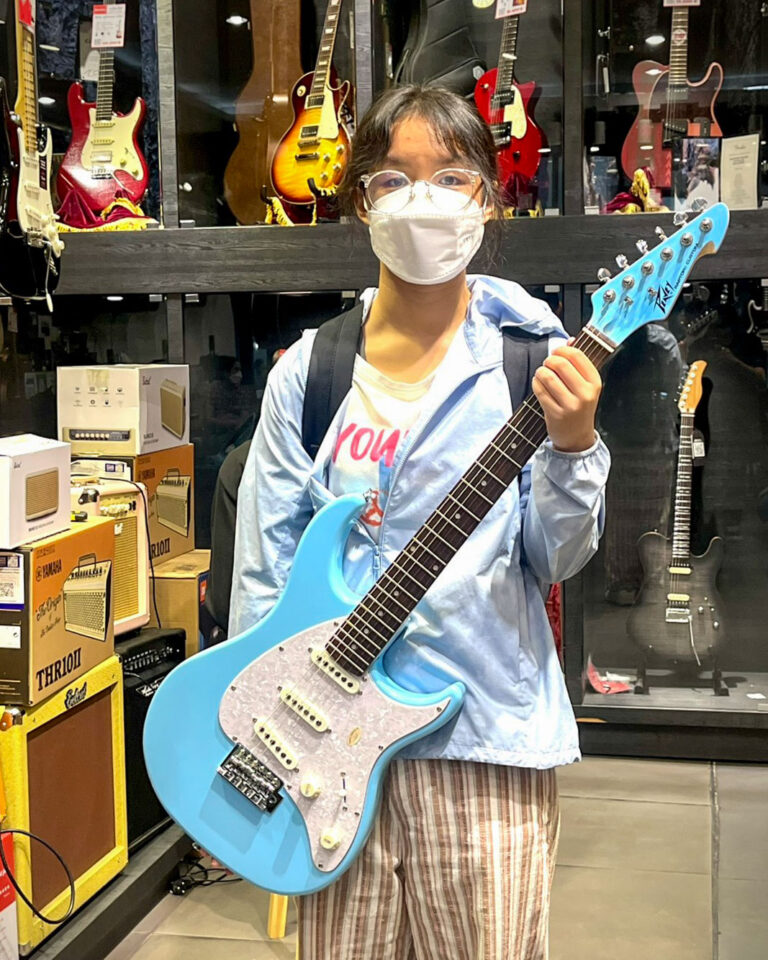 Person wearing a white face mask and glasses holds a light-blue electric guitar in a guitar shop, with guitars on display behind.