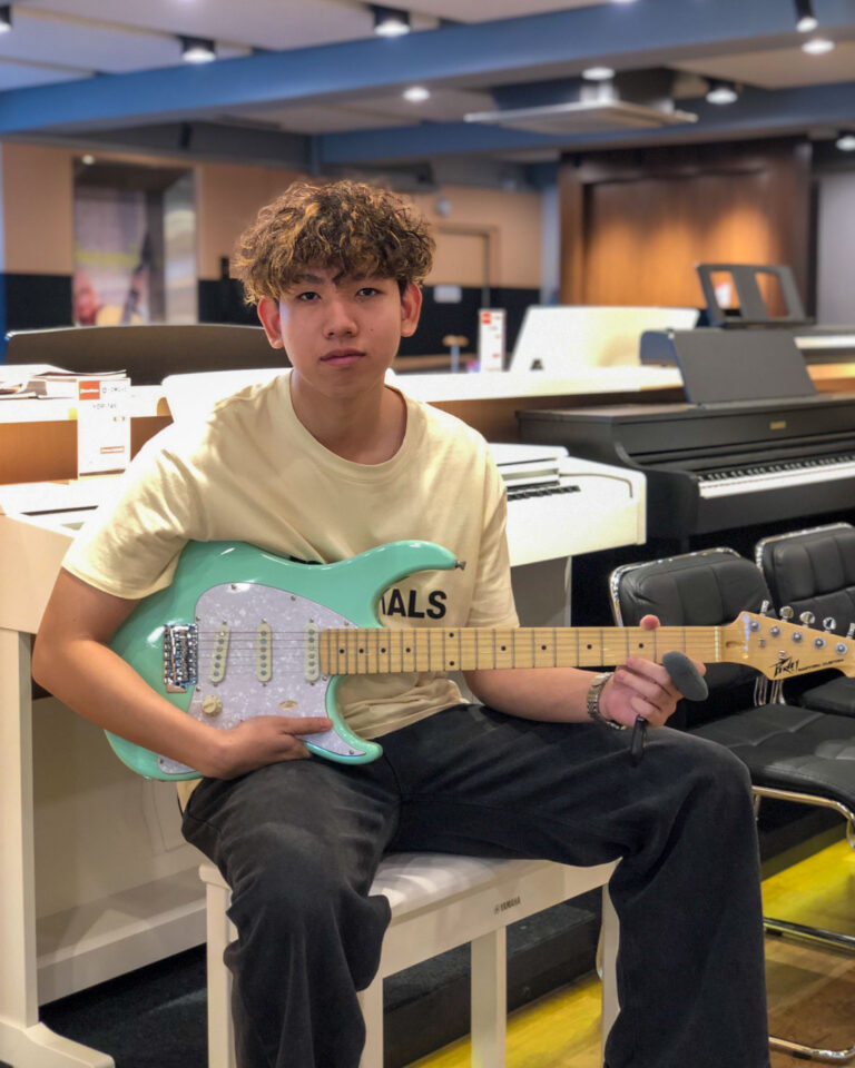 Young man sits in a music store holding a turquoise electric guitar, with keyboards and instruments in the background.