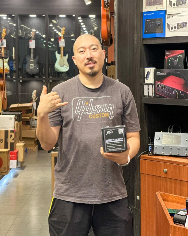 Man in a gray Gibson Custom shirt stands in a guitar shop, holding a boxed item labeled 'Session-LTD' and making a shaka gesture. Behind him are glass cases with guitars and gear.