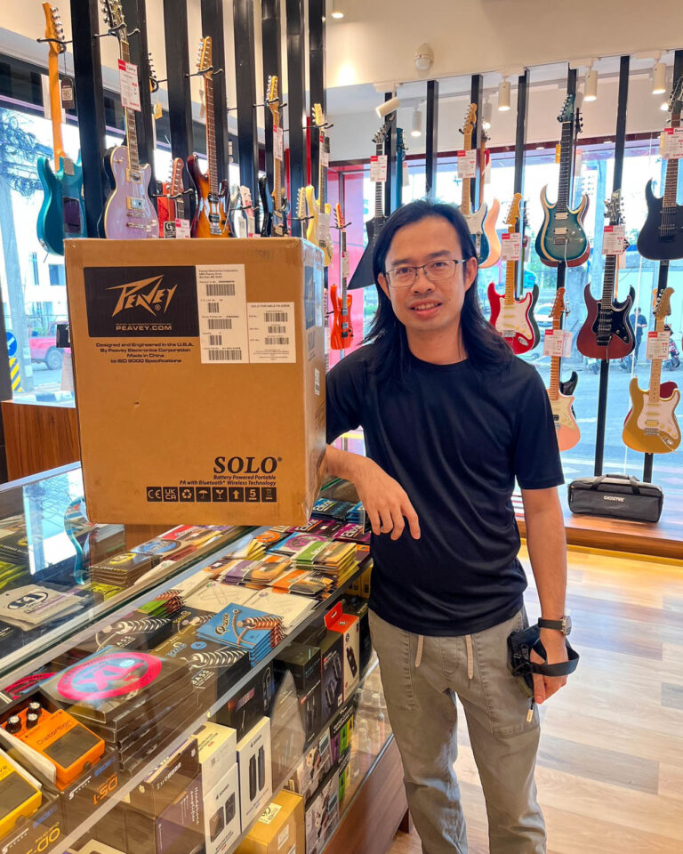 Man wearing a black shirt stands next to a large cardboard shipping box in a guitar shop, with guitars hanging on the wall behind.