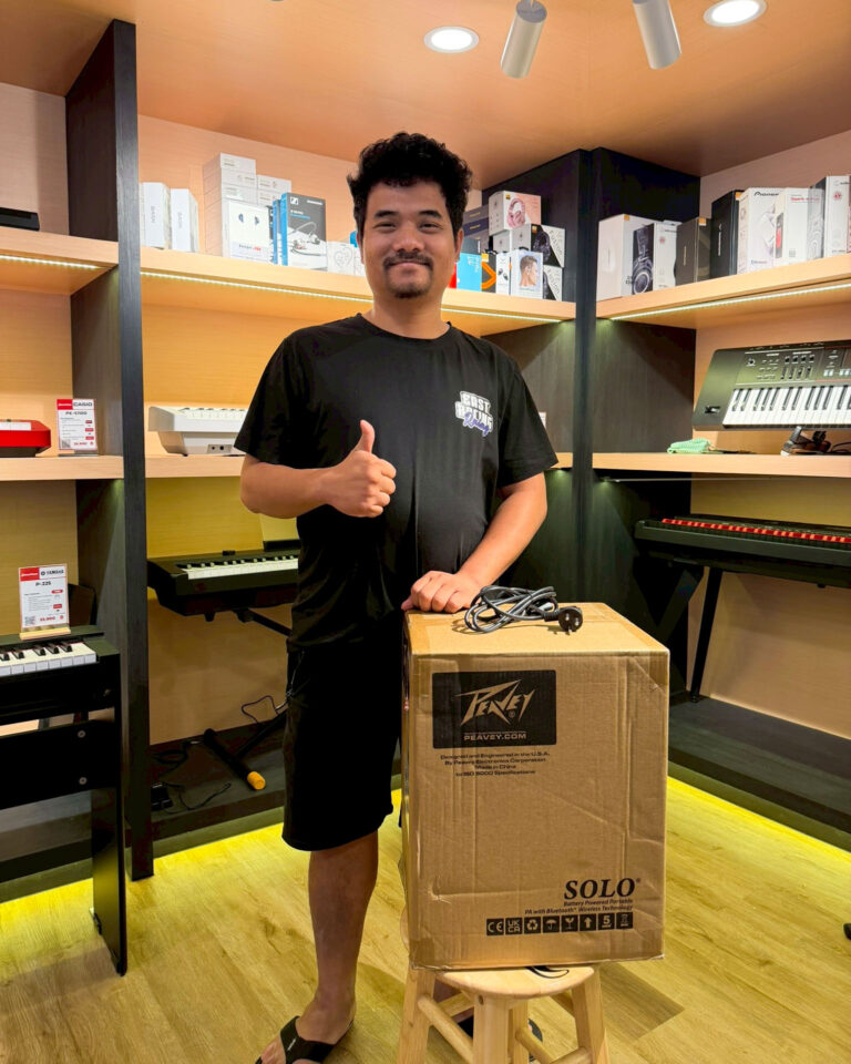 Smiling man in a black T-shirt gives a thumbs up beside a large cardboard box labeled SOLO in a music store, with keyboards and audio gear on shelves behind him.