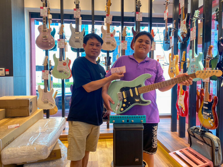 Two men in a guitar shop pose with a mint-green electric guitar and a blue amplifier beside them.