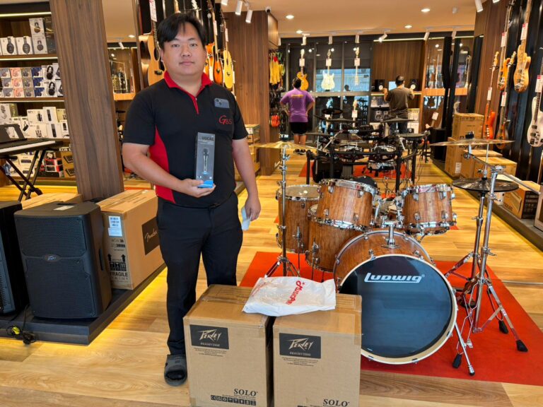 Salesperson in a music store stands beside a Ludwig drum kit, holding a microphone box with boxed gear around him.