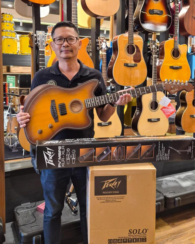 Man in a guitar shop holding a sunburst acoustic guitar with many guitars on display in the background.