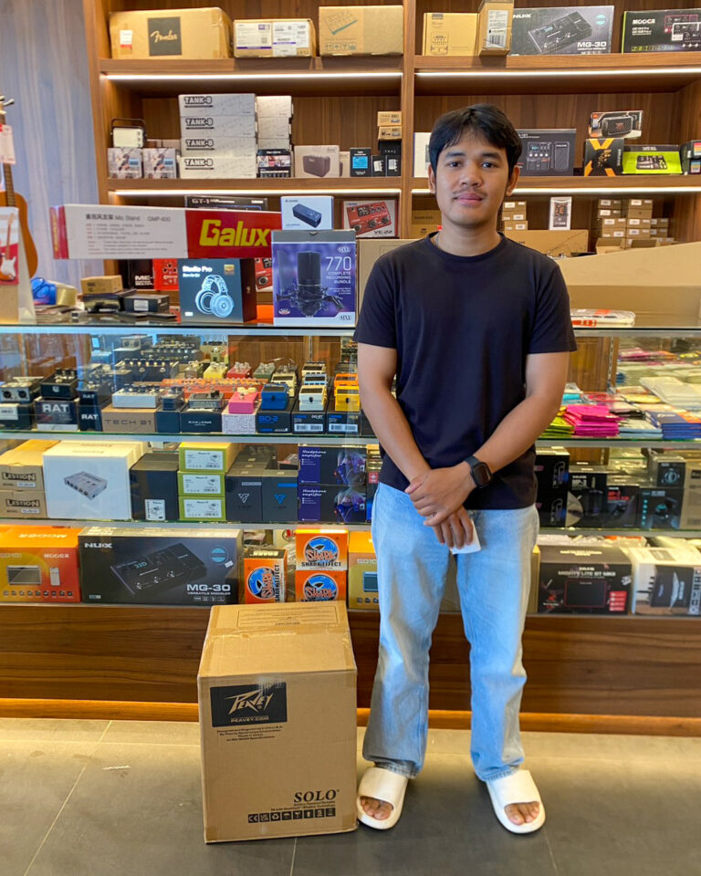 Young man in a black T-shirt standing in an electronics/music store beside a large cardboard box labeled SOLO, shelves of audio gear behind him.