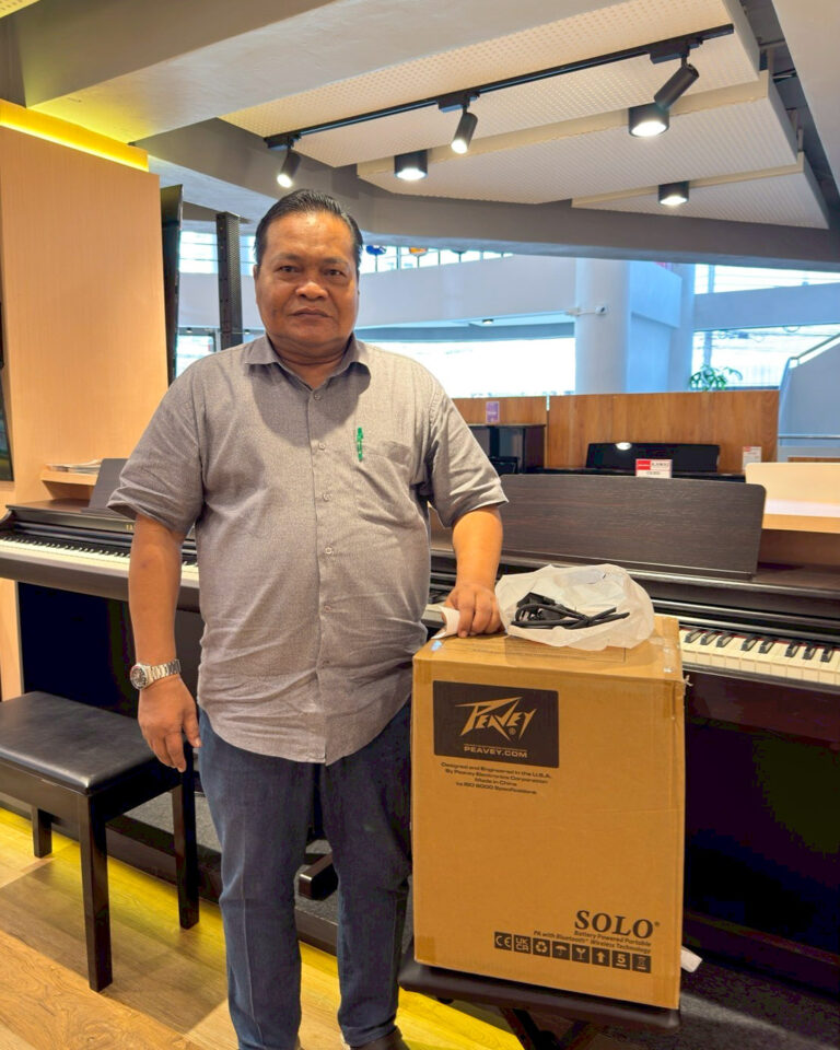 Man in a gray shirt stands beside a piano in a showroom, with a large SOLO box on a bench nearby.