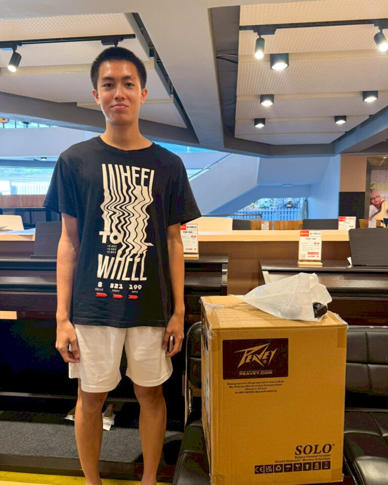 Young man wearing a black graphic t-shirt and white shorts stands next to a large cardboard box labeled SOLO in a showroom.