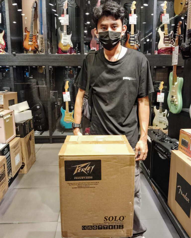 Man wearing a black t-shirt and mask stands behind a large cardboard box in a guitar store.