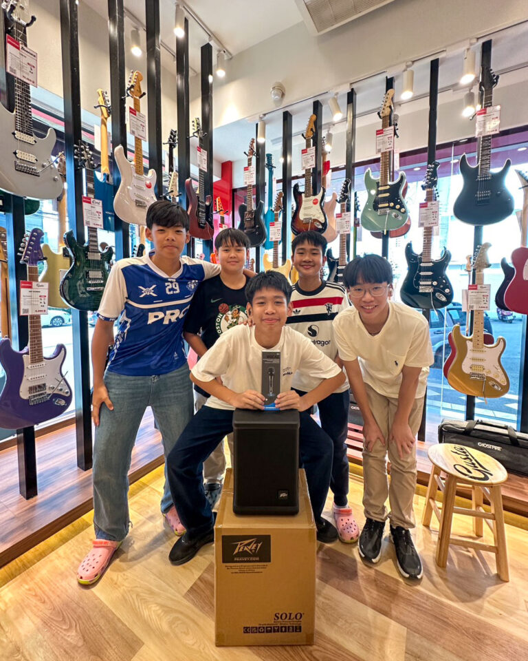 Five boys in a guitar shop pose for a photo among hanging electric guitars; one sits on a box holding a boxed microphone.