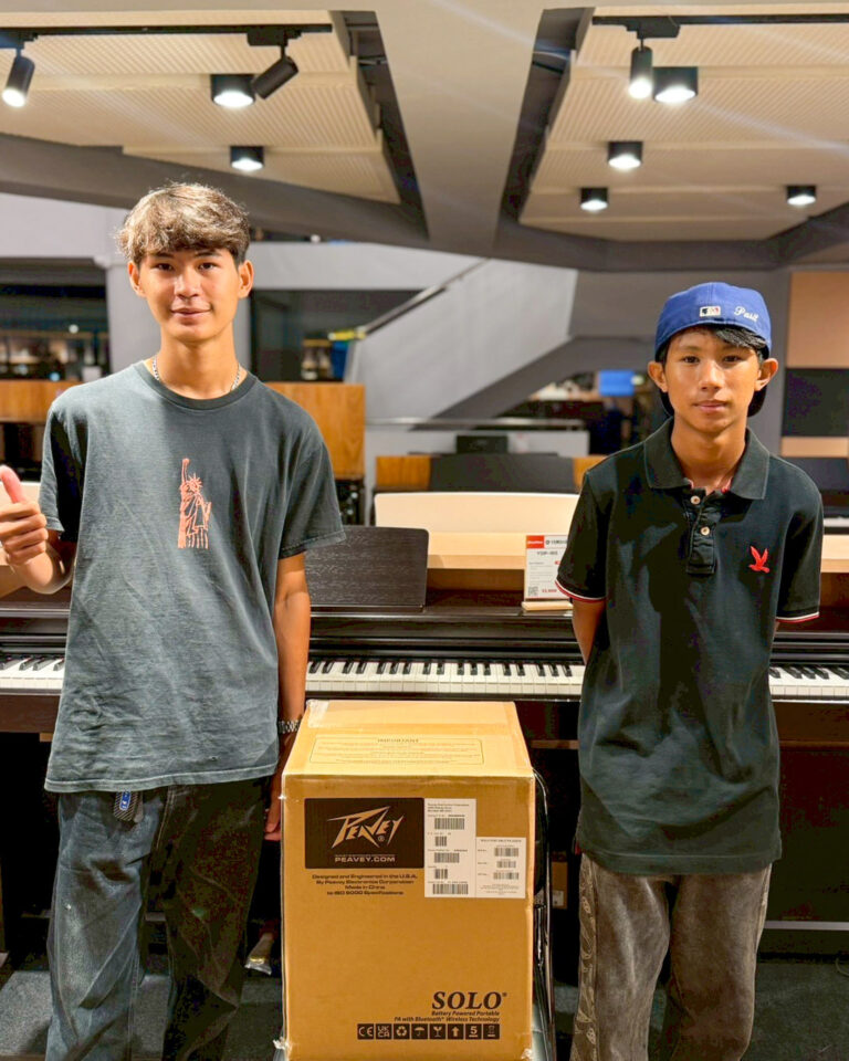 Two young men stand beside a large cardboard box in a music store, with a piano behind them, looking at the camera.