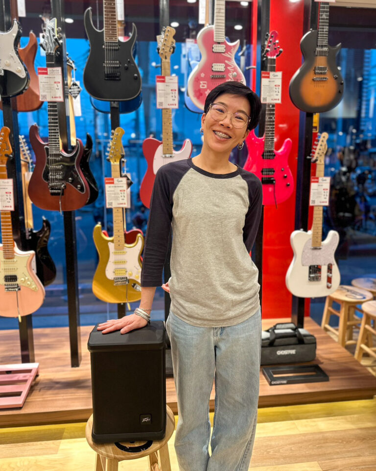 Smiling woman with glasses leaning on a black speaker in a guitar shop, colorful electric guitars on display behind her.