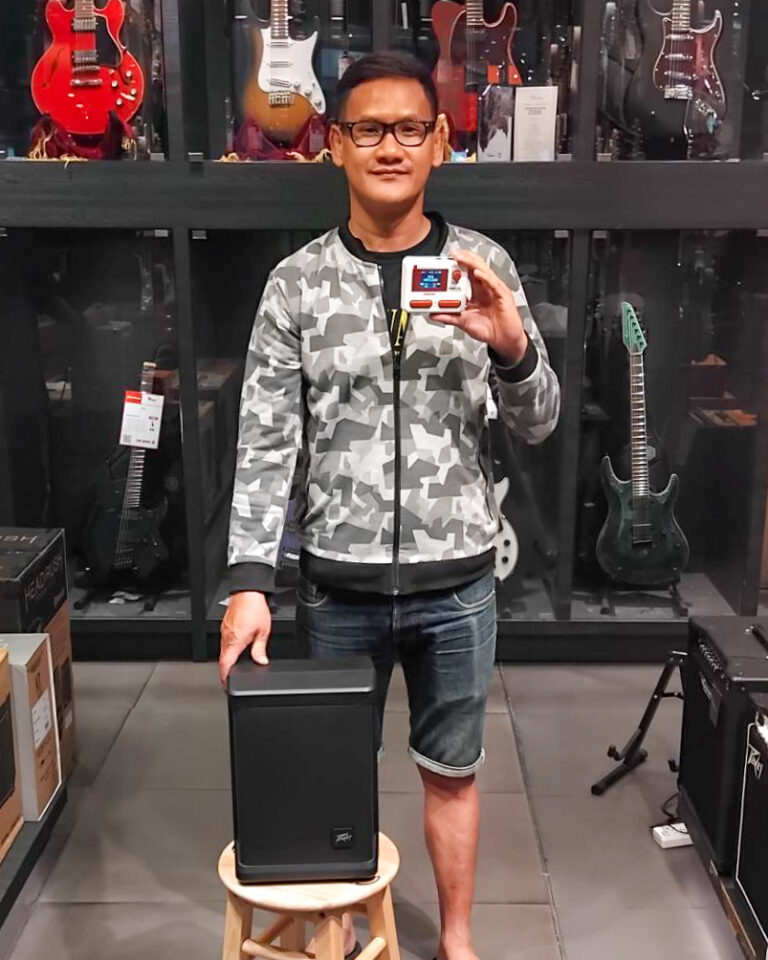 Person in a music store posing with a small red-and-white handheld device in front of a large black speaker on a stool, guitars visible in the background