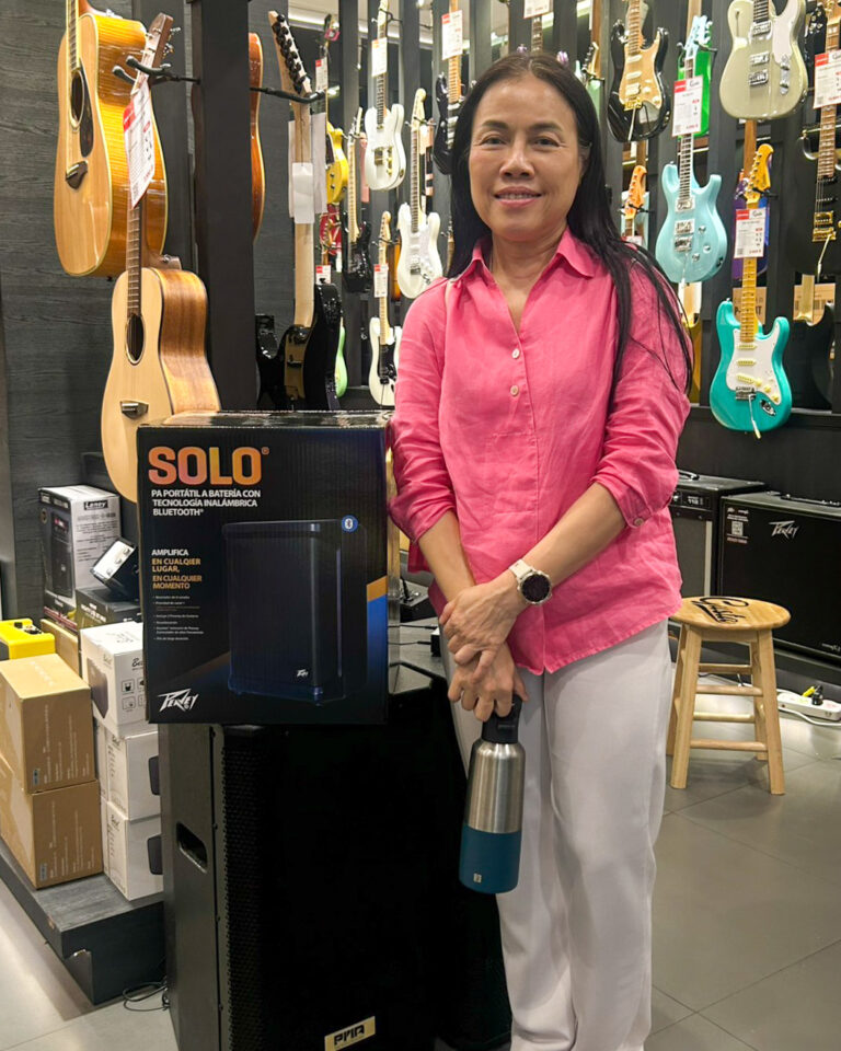 Woman in pink blouse smiles in a guitar shop, standing beside a SOLO portable speaker box.