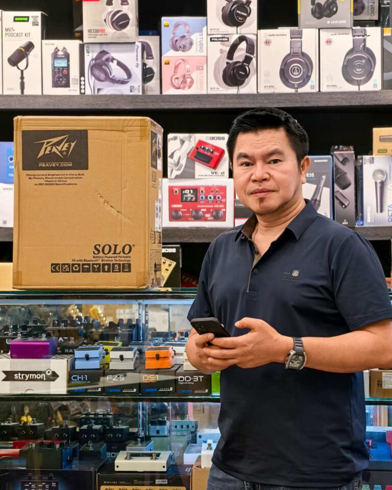 Man in a dark polo shirt holds a smartphone beside a large Solo speaker box in an audio gear shop.