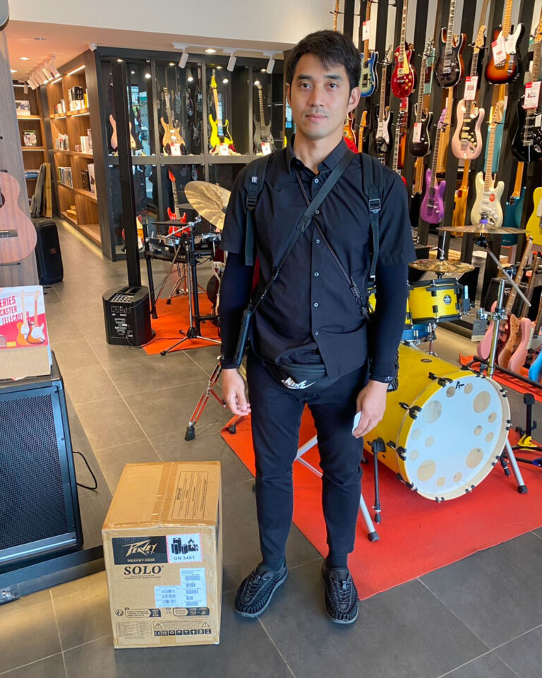 Man in black outfit with crossbody bag standing in a guitar store, shelves of colorful guitars and a drum set visible behind him, with a cardboard box on the floor.