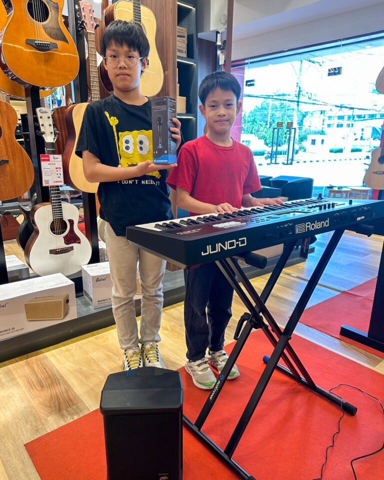 Two kids stand by a Roland keyboard in a guitar shop; one holds a mic box while the other plays the keys nearby.