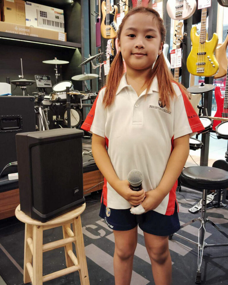 Young girl in a white polo with red panels holding a microphone in a music shop, with drums and guitars on display in the background.