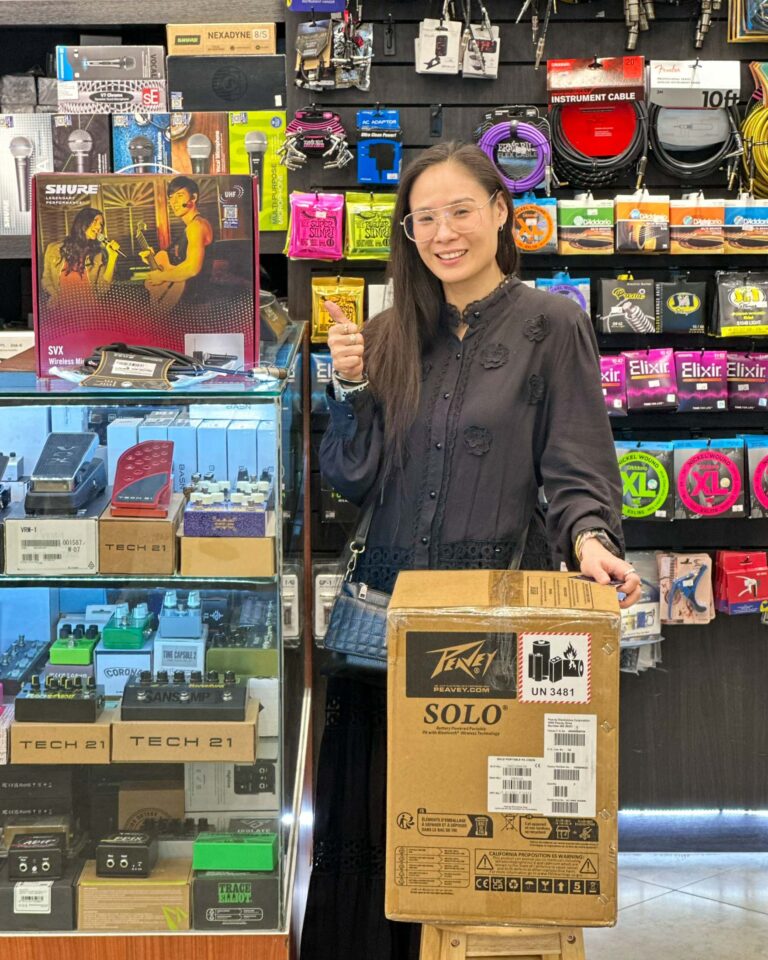 Smiling woman in a music gear shop giving a thumbs up, surrounded by cables, pedals, and boxed gear on display behind her.