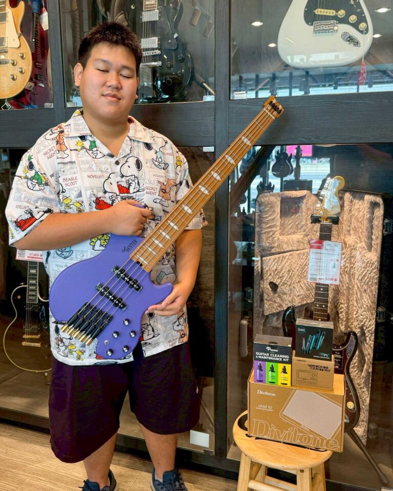 Young person in a music store posing with a purple bass guitar in front of guitar displays behind glass cabinets.