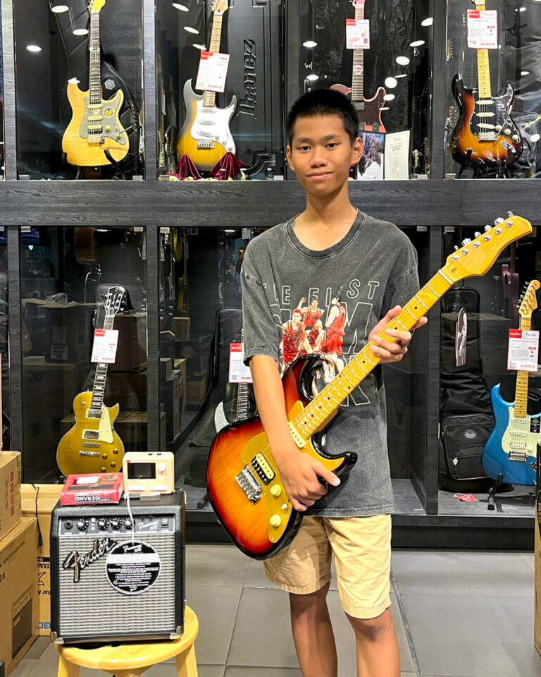Young man in a guitar shop posing with a sunburst electric guitar in his hands, shelves of guitars behind him.