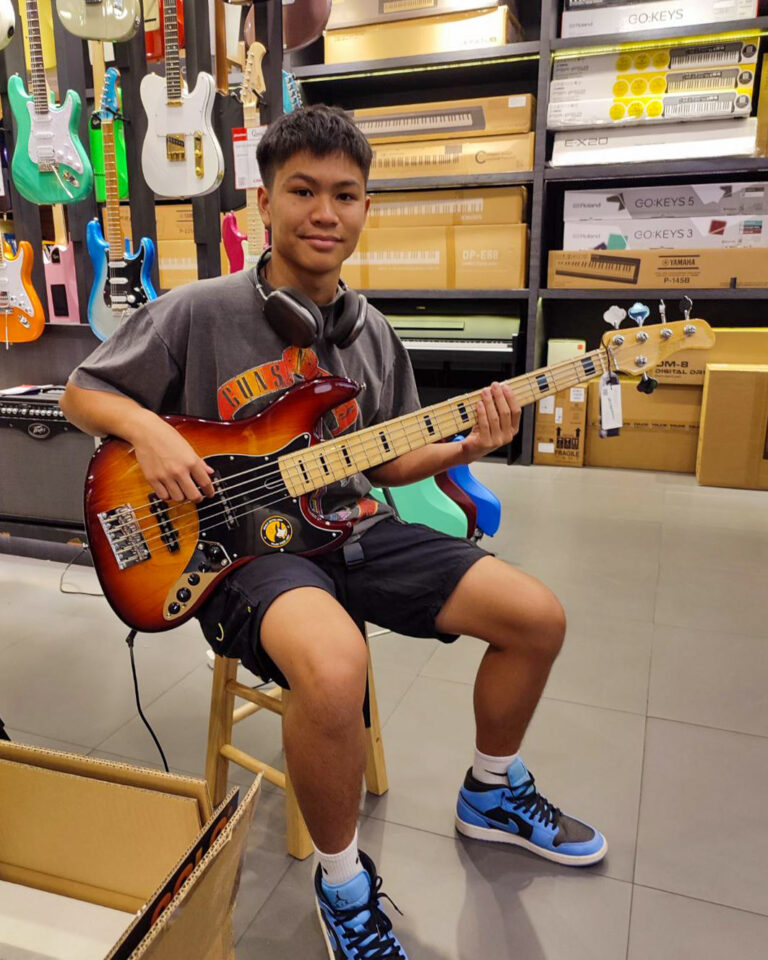 Young man sits on a stool in a music store, playing a sunburst bass guitar with headphones around his neck in the aisle of instruments and boxes behind him