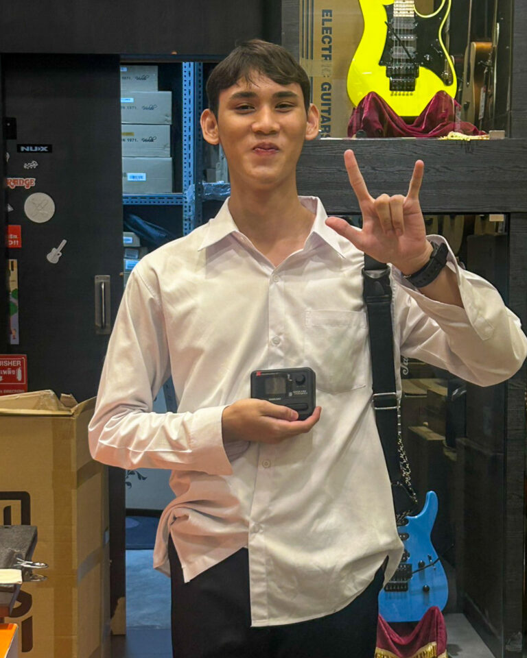 Young man in a white shirt holds a small action camera and makes the 'rock on' sign in a music-store setting with guitars in the background.