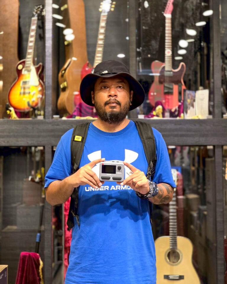 Man wearing a blue Under Armour shirt and backpack stands in a guitar shop, holding a small action camera toward the camera with neutral expression.