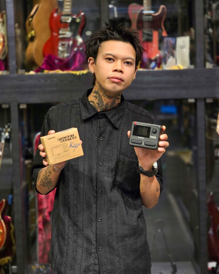 Person with tattoos wearing a black shirt holding a boxed gadget and a handheld device in a storefront window with guitars on display behind him