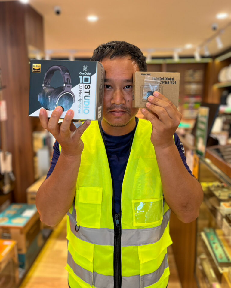 Man in a high-visibility vest holding two boxed products in a store: headphones box in left hand and compact gadget box in right hand.