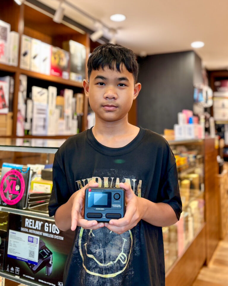 Boy in a shop holding a small blue compact camera toward the viewer, smiling slightly.