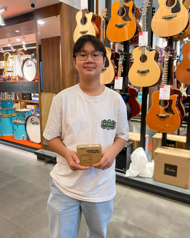 Young boy wearing glasses and a white T-shirt stands in a guitar shop holding a small brown box beside a display of guitars.