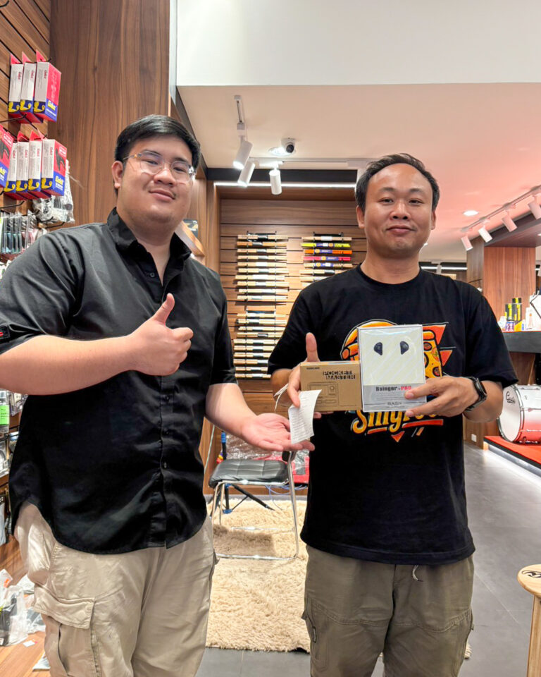 Two men in a music shop pose for a photo; one gives a thumbs up while the other holds two boxed audio items. Shelves with instrument accessories in the background.