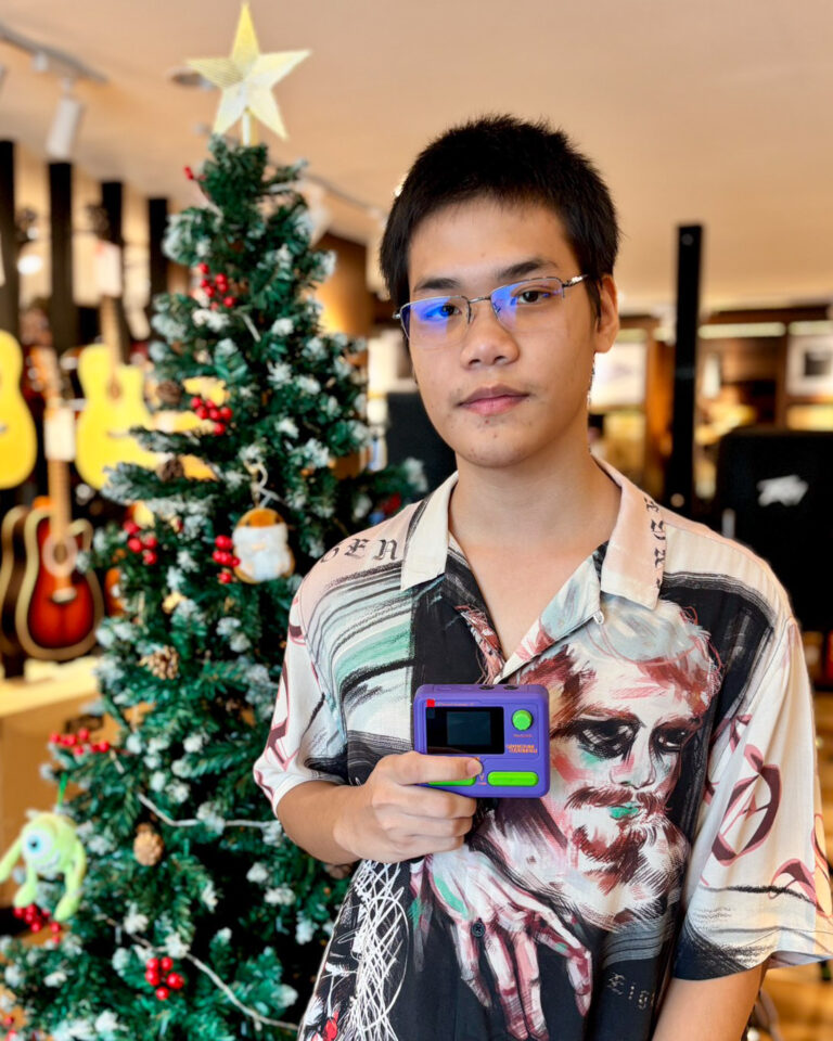 Young man with glasses and a patterned shirt holds a purple handheld game console in a guitar shop decorated for Christmas.