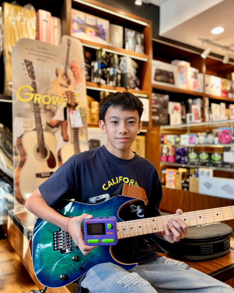 Boy in a music store holds a blue electric guitar and a small purple handheld game console, looking at the camera.
