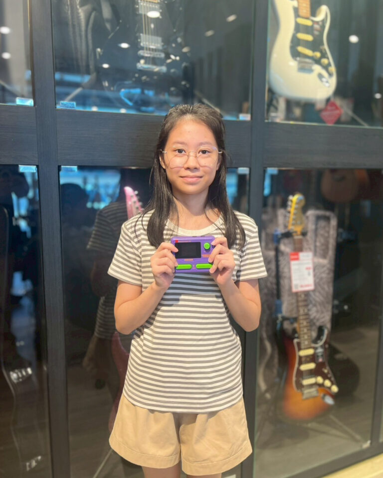 Girl with glasses in a striped shirt and beige shorts holding a purple-and-green handheld game console in a guitar store window display behind her.