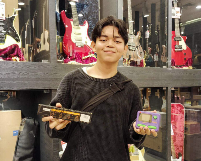 Young man in a guitar shop holds a chocolate bar package in one hand and a purple handheld gaming device in the other.