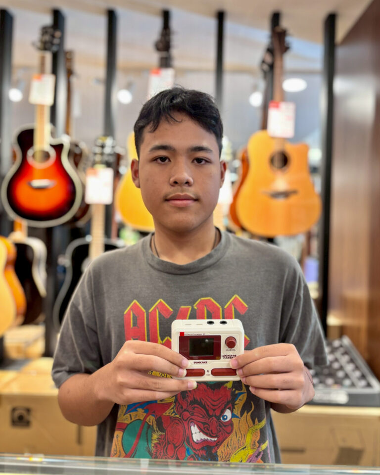 Young man in a music store holding a white retro handheld game console, with guitars blurred in the background