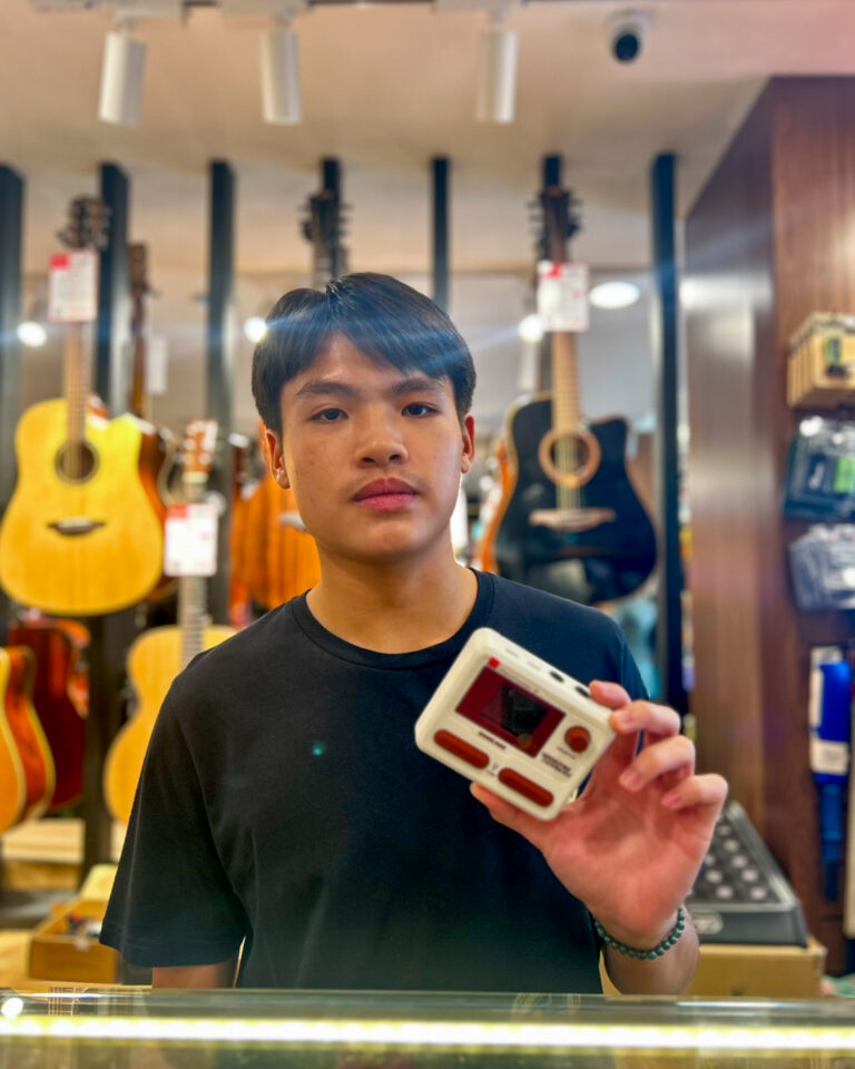 Young man in a guitar shop holding a white handheld game console toward the camera.