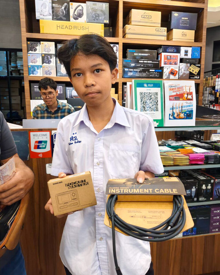 Young man in a white shirt standing behind a counter in an electronics/music store, holding a boxed instrument cable and another boxed accessory while shelves of audio gear loom in the background.