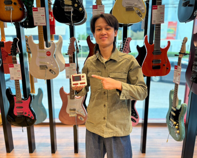 Person in a guitar shop holding a small white handheld guitar tuner and pointing at it toward the camera.