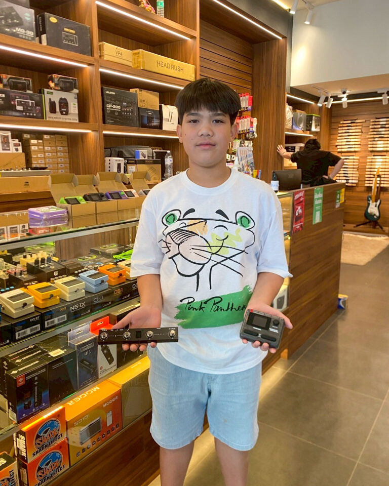 Young boy in a music store holding two guitar effects pedals in front of shelves of audio gear.