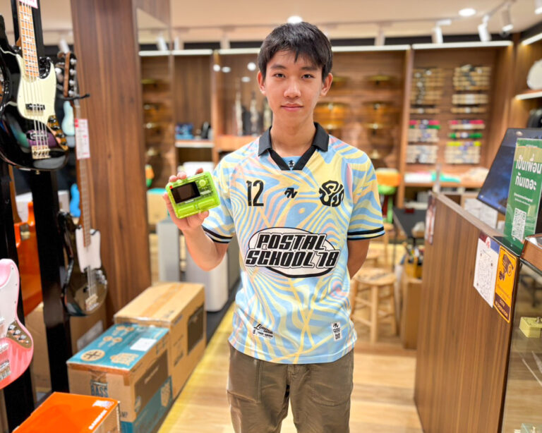 Teen boy in a light blue jersey holds a bright green handheld device in a guitar shop with guitars on display behind him.