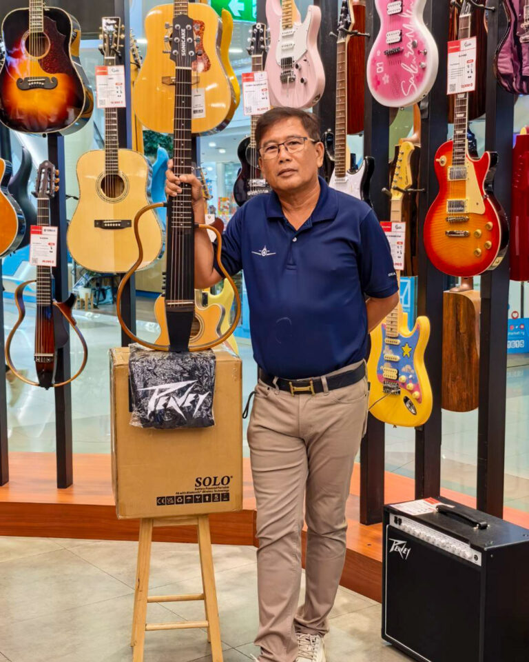 Man in a blue polo shirt stands in a guitar shop, holding an acoustic guitar among a wall of colorful guitars on display.