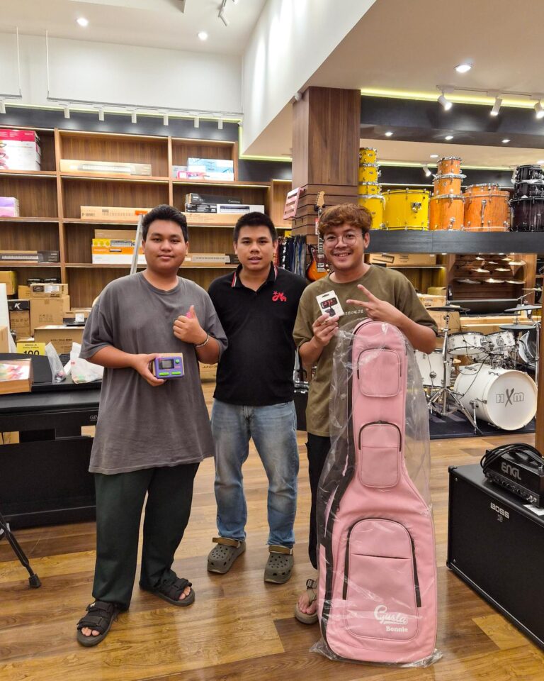 Three young men pose in a music store with shelves of boxes and instruments in the background; one holds a handheld device, another wears a black polo, the third has a pink wrapped guitar case nearby.