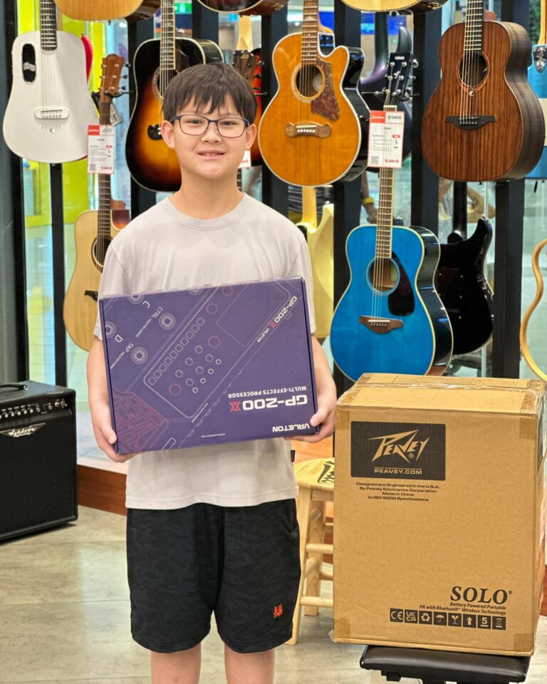 Smiling boy with glasses in a guitar shop holding a large purple box, with guitars on display in the background.