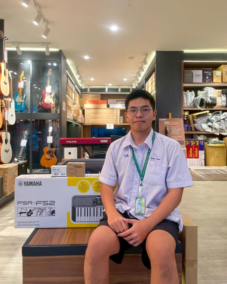 Young man wearing a lanyard sits in a music store beside a Yamaha PSR-F52 keyboard box, with guitars on display behind him.