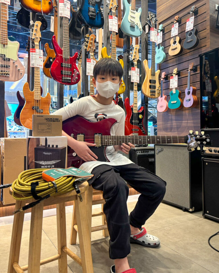 Boy wearing a mask sits in a guitar shop holding a red electric guitar with many guitars on display behind him.