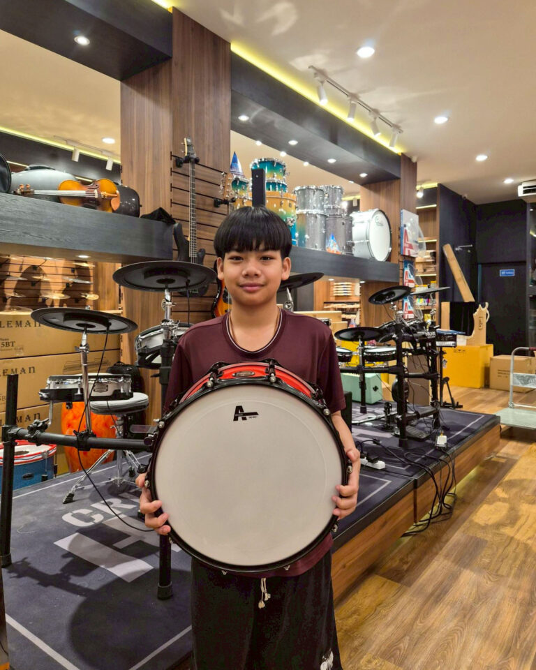 Boy standing in a music store, holding a large bass drum and smiling, with drums and guitars displayed behind him.