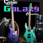Stage setup featuring two colorful electric guitars (purple sparkle and teal) in front of a drum kit, with a large Gusta backdrop logo behind them for a music performance. ลดราคาพิเศษ